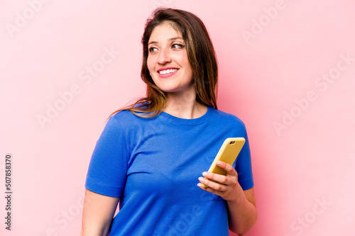 Young caucasian woman holding mobile phone isolated on pink background looks aside smiling  cheerful and pleasant.
