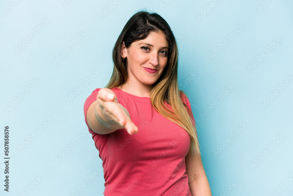 Young caucasian woman isolated on blue background stretching hand at camera in greeting gesture. Young caucasian woman isolated on blue background stretching hand at camera in greeting gesture.