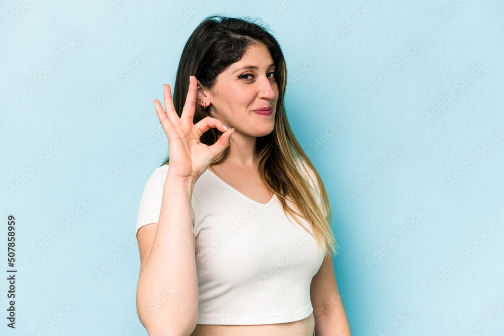 Young caucasian woman isolated on blue background winks an eye and holds an okay gesture with hand. Young caucasian woman isolated on blue background winks an eye and holds an okay gesture with hand.