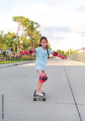 Asian little girl child skateboarder wearing safety and protective equipment playing on skateboard. Kid skateboarding on the road outdoors