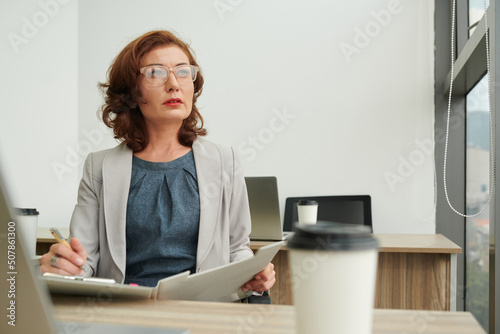 Pensive middle-aged female entrepreneur in glasses filling documant in folder photo