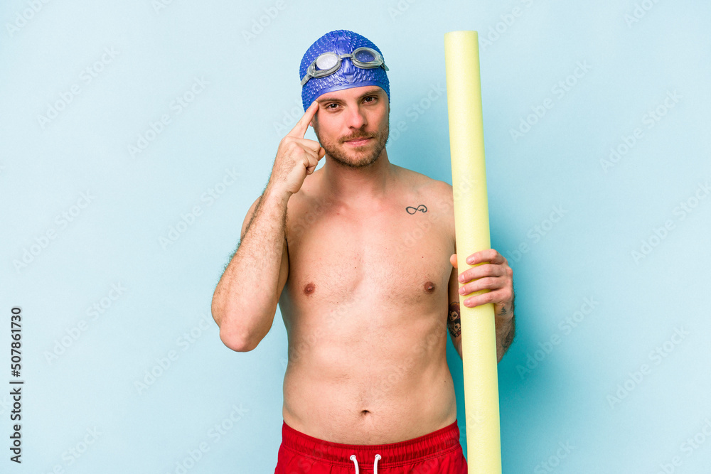 Young caucasian swimmer man holding foam stick isolated on blue background pointing temple with finger, thinking, focused on a task.