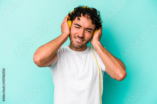 Young caucasian man listening to music isolated on blue background covering ears with hands.
