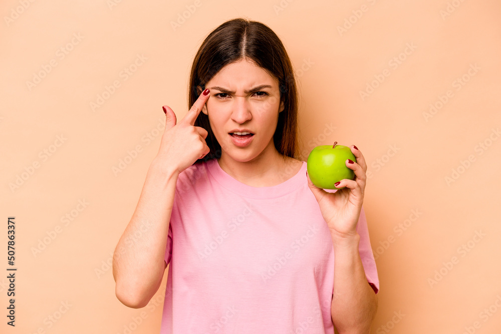 Young hispanic woman holding an apple isolated on beige background showing a disappointment gesture with forefinger. Young hispanic woman holding an apple isolated on beige background showing a disappointment gesture with forefinger.