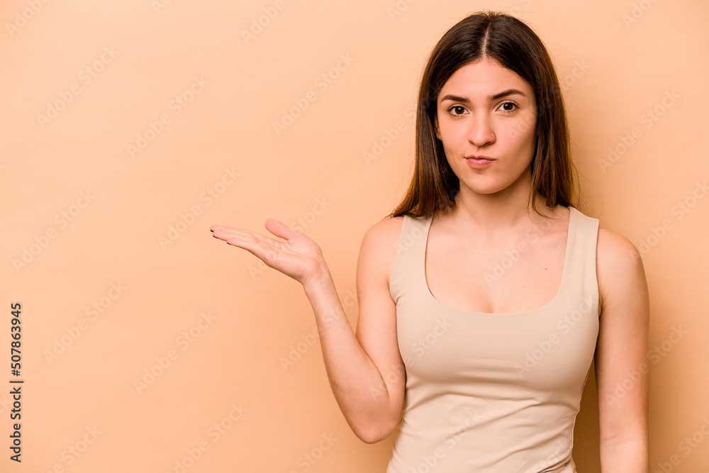 Obraz premium Young hispanic woman isolated on beige background showing a copy space on a palm and holding another hand on waist.
