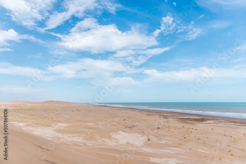 Dunes in front of the beach with the blue sky in the background and the horizon in the sea with some clouds.