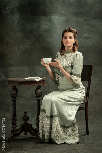 Vintage portrait of young beautiful girl in gray dress of medieval style drinking tea isolated on dark background. Comparison of eras concept, flemish style. Art, beauty