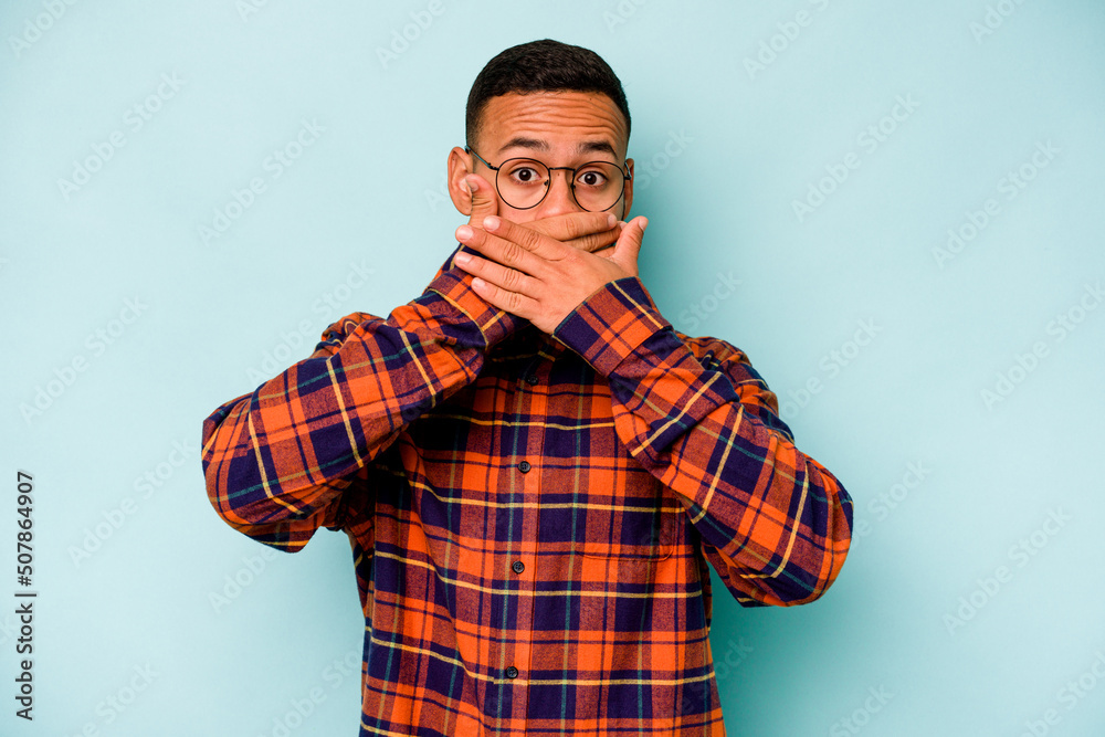 Young hispanic man isolated on blue background covering mouth with hands looking worried.