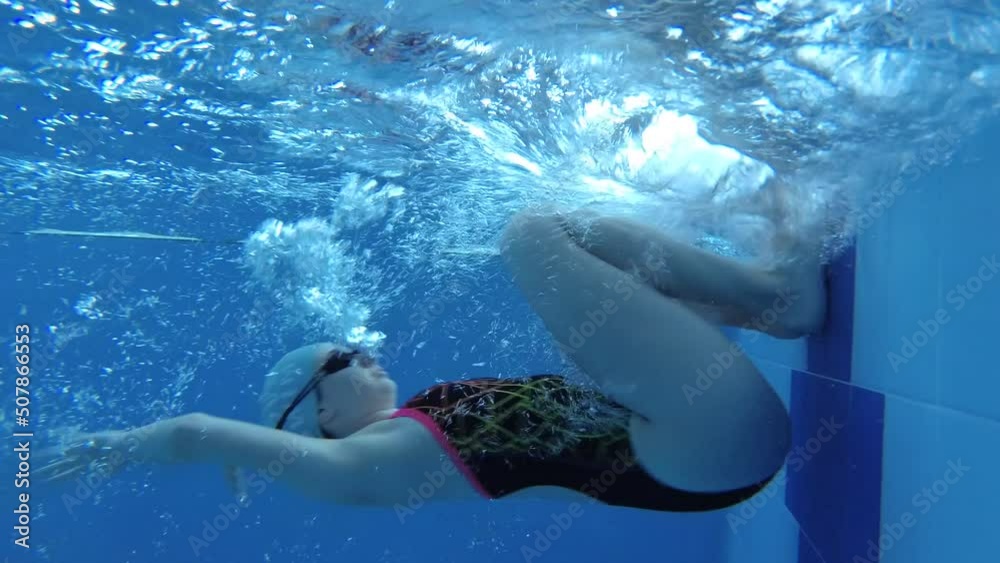 sportswoman swimmer in a tracksuit, in diving goggles, in a swimming cap is training in the pool. The athlete swims underwater. Underwater shooting, wide angle, 4k
