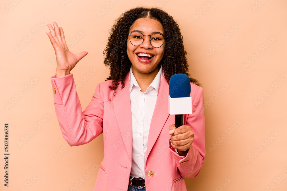 Young African American TV presenter woman isolated on beige background receiving a pleasant surprise, excited and raising hands. - obrazy, fototapety, plakaty 