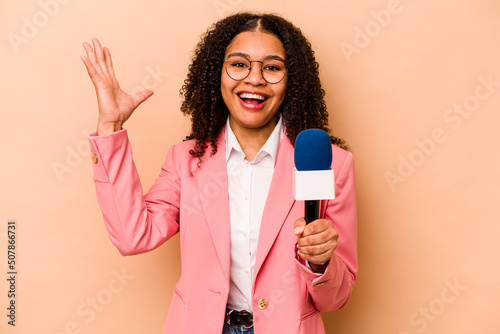 Young African American TV presenter woman isolated on beige background receiving a pleasant surprise, excited and raising hands.