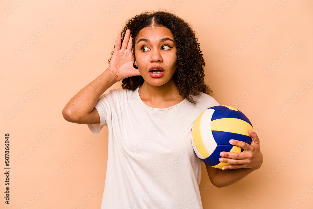 Young African American woman playing volleyball isolated on beige background trying to listening a gossip.
