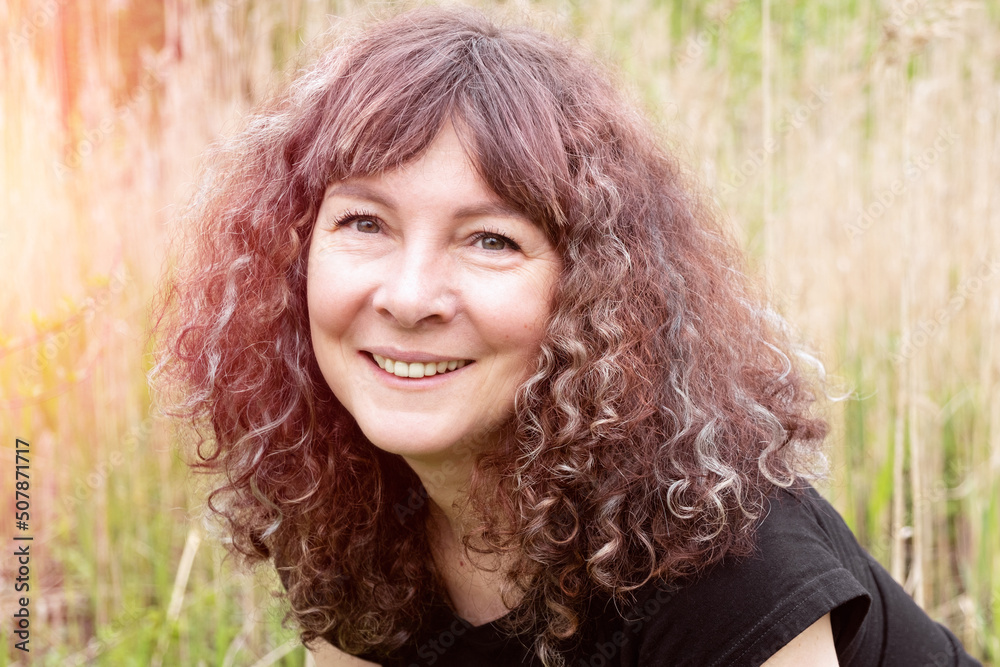 Portrait of beautiful caucasian white woman with brown curly hair looking at camera and smiling. Happy woman on the nature on vacation. Close up. Blurred background. Portrait of beautiful caucasian white woman with brown curly hair looking at camera and smiling. Happy woman on the nature on vacation. Close up. Blurred background.