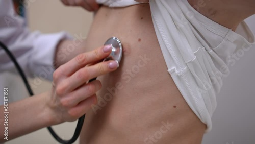 Close-up listening auscultation with stethoscope in pediatric clinic indoors. Unrecognizable Caucasian boy raising T-shirt as doctor using equipment diagnosing illness in hospital photo