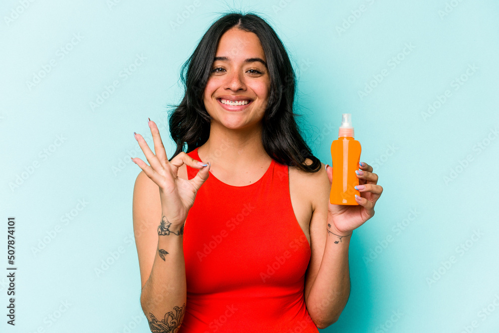 Young hispanic woman holding sun cream isolated on blue background cheerful and confident showing ok gesture. Young hispanic woman holding sun cream isolated on blue background cheerful and confident showing ok gesture.