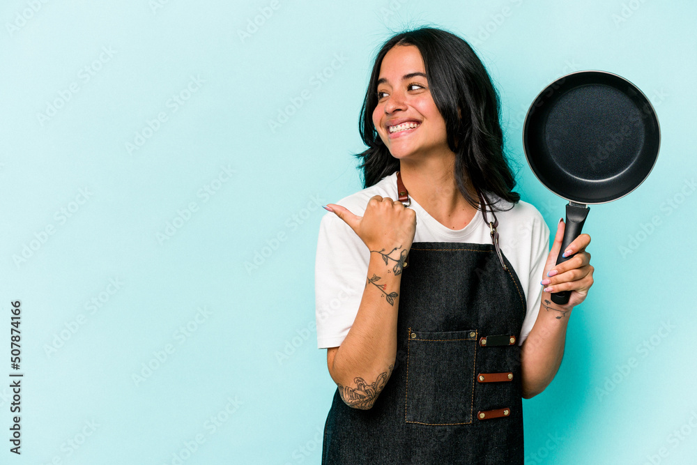 Young hispanic cooker woman holding frying pan isolated on blue background points with thumb finger away, laughing and carefree. Young hispanic cooker woman holding frying pan isolated on blue background points with thumb finger away, laughing and carefree.