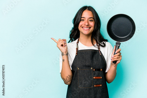 Young hispanic cooker woman holding frying pan isolated on blue background smiling and pointing aside, showing something at blank space.