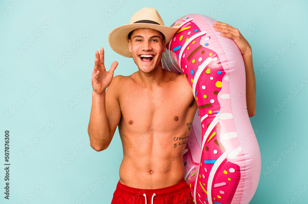Young caucasian man holding an inflatable donut isolated on blue background receiving a pleasant surprise, excited and raising hands.