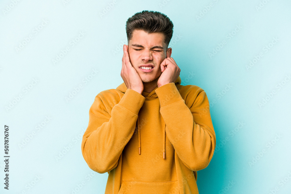 Young caucasian man isolated on blue background covering ears with fingers, stressed and desperate by a loudly ambient. Young caucasian man isolated on blue background covering ears with fingers, stressed and desperate by a loudly ambient.