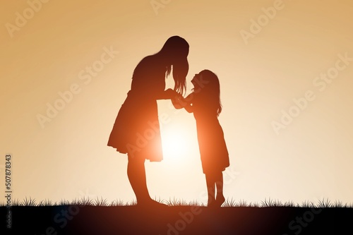 Silhouette of a young mother lovingly holding hands with her happy little child in front of a sunset in the sky.