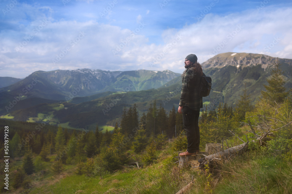 A lone man in a flannel shirt with a backpack on his back stands on a nature trail in the mountain landscape of Austria, with the Schneealpe and Raxalpe mountain ranges visible in the background.