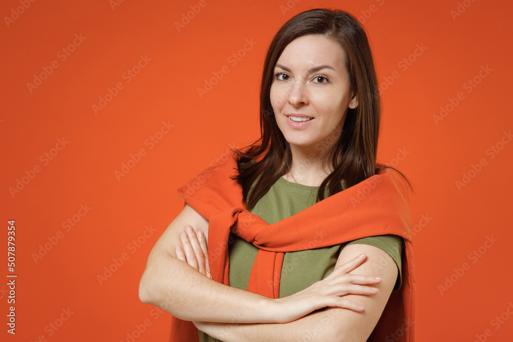 Obraz premium Young smiling happy fun woman 20s wearing khaki t-shirt tied sweater on shoulders look camera hold hands crossed folded isolated on plain orange background studio portrait. People lifestyle concept.