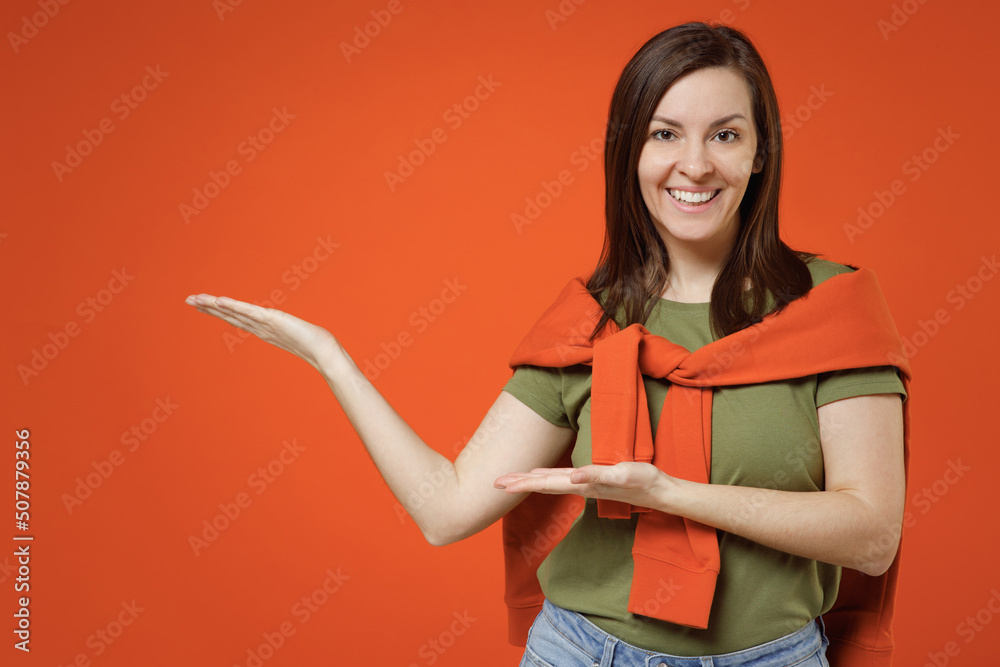 Young smiling fun happy woman 20s wearing khaki t-shirt tied sweater on shoulders point hands arms aside on workspace area mock up isolated on plain orange background studio. People lifestyle concept.