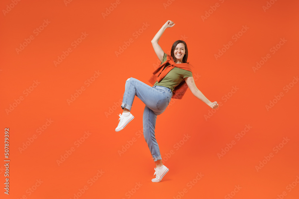 Full body young excited fun happy woman 20s wearing khaki t-shirt tied sweater on shoulders doing winner gesture celebrate clenching fists say yes isolated on plain orange background studio portrait
