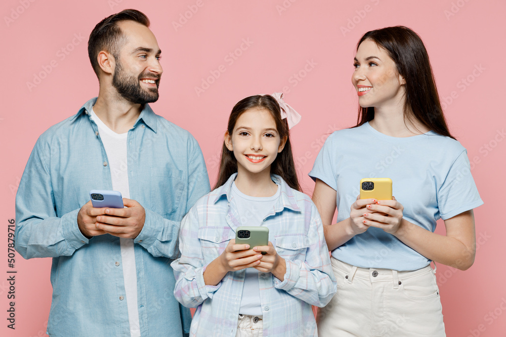 Young smiling fun parents mom dad with child kid daughter teen girl in blue clothes hold in hand use mobile cell phone look to each other isolated on plain pastel pink background. Family day concept Young smiling fun parents mom dad with child kid daughter teen girl in blue clothes hold in hand use mobile cell phone look to each other isolated on plain pastel pink background. Family day concept