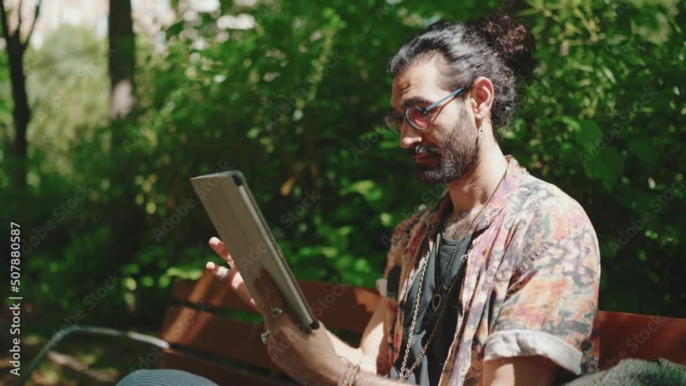 Concentrated curly-haired bearded man in eyeglasses looking at tablet while sitting on the bench outdoors Concentrated curly-haired bearded man in eyeglasses looking at tablet while sitting on the bench outdoors