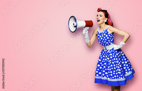 Purple haired woman holding red megaphone, shout advertising something. Girl in blue pin up style with mega phone loudspeaker. Rose pink background with mock up. Female model in retro fashion dress.