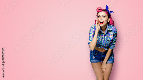 Happy excited woman holds hand near open mouth, looking aside and saying something. Girl dressed in pinup style. Red purple haired pin up model at retro fashion vintage concept, pink background.