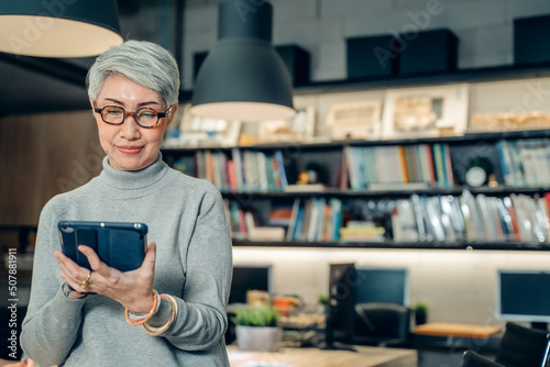 Senior confident business working woman, mentor, teacher with white short hair and glasses stand and use tablet, Portrait mature female middle aged in office with colleagues discuss work at background
