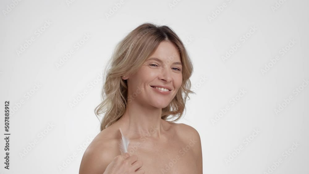 Beautiful happy middle aged lady caressing her smooth perfect skin with white feather, enjoying her body condition