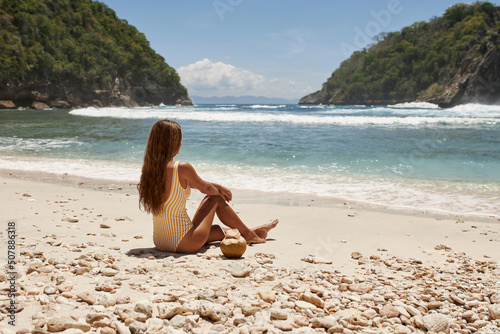 Beautiful model in a yellow swimsuit is drinking a coconut on the beach. Rear view of a woman in a bikini sitting near the ocean with a coconut.