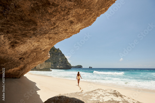 Young beautiful girl in a yellow swimsuit is sunbathing while standing on a tropical beach with white sand and turquoise water. Vacation on Diamond Beach in Nusa Penida Bali Indonesia photo