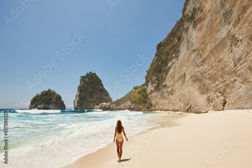 Young beautiful girl in a yellow swimsuit is sunbathing while standing on a tropical beach with white sand and turquoise water. Vacation on Diamond Beach in Nusa Penida Bali Indonesia