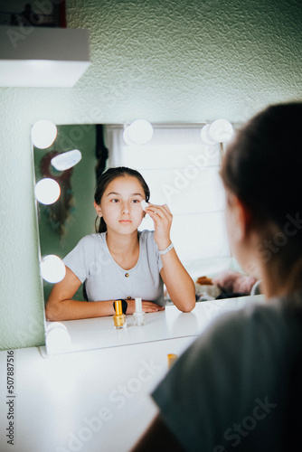 Chica joven sentada en su habitación cuidándose la piel frente a su espejo con luces. photo