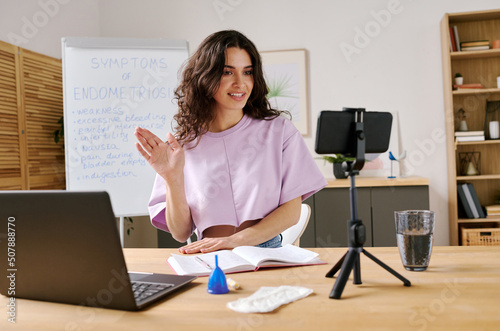 Fototapeta Young woman sitting at desk in front of smartphone on tripod starting live strea