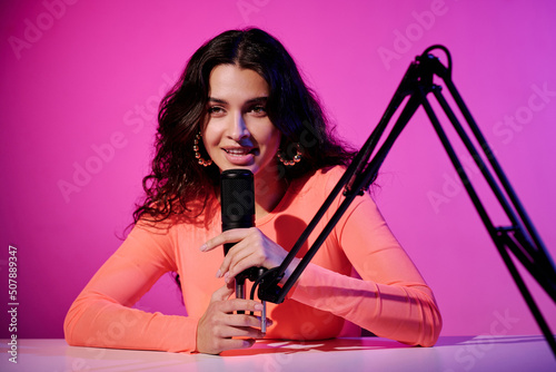 Beautiful young female influencer sitting at desk in studio in pink neon light whispering in microphone for ASMR video