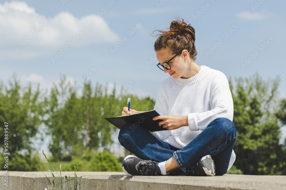 Student woman in sweater and glasses sitting outdoors and writing in her diary.