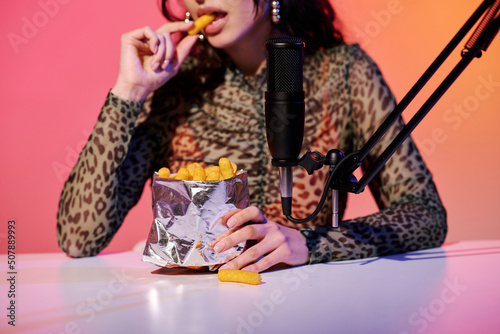 Stylish young female blogger sitting in studio in pink and yellow neon light recording corn puff snacks eating sounds for ASMR vlog