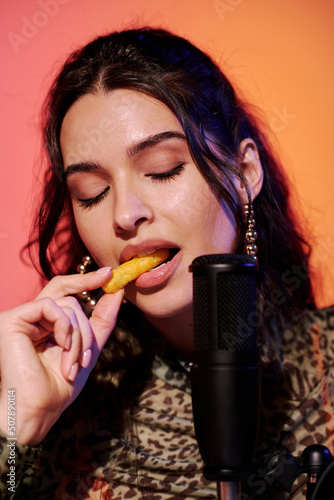 Attractive young woman with eyes closed sitting in studio in pink and yellow neon light recording corn puff snacks eating sounds for ASMR vlog