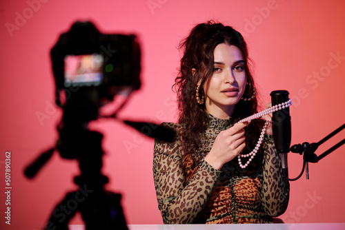 Stylish young Caucasian woman sitting in studio in pink neon light recording sounds and video for ASMR vlog using bead necklace photo