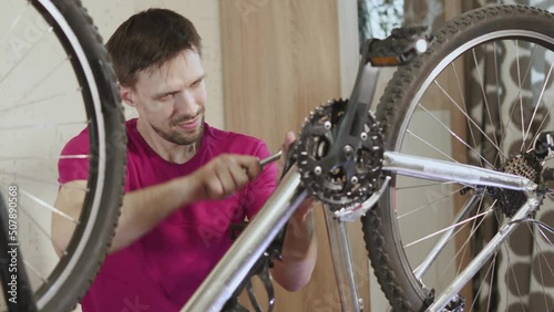 Mechanic tightens bolt with socket wrench on bicycle bottom bracket shaft.. Man turns chainring system, part of transmission, checks repair of bicycle. Guy is happy with work, smiling, slow motion. photo