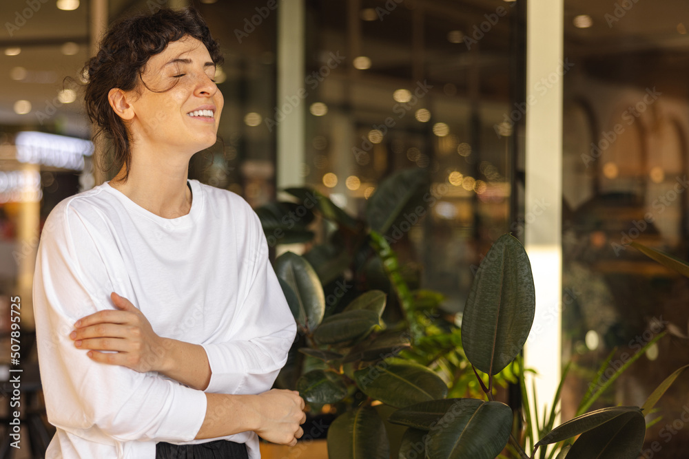 Cute young caucasian girl smiles with eyes closed and hugs herself indoors with plants. Brunette wears white sweatshirt enjoying her vacation. Modern lifestyle, people and youth concept Cute young caucasian girl smiles with eyes closed and hugs herself indoors with plants. Brunette wears white sweatshirt enjoying her vacation. Modern lifestyle, people and youth concept