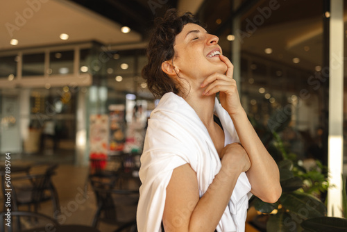 Happy young caucasian woman laughs with eyes closed and head tilted back standing indoors. Brunette is wearing white sweatshirt over her shoulders. Concept of enjoying moments