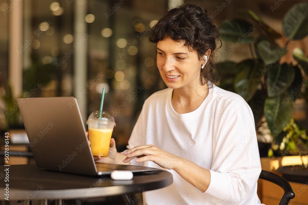 Positive young caucasian girl uses her laptop sitting in cafe. Brunette is chatting on social networks with friends and drinking juice. Concept of technology 