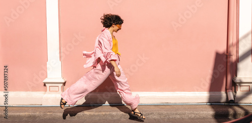 Full-length, young, stylish caucasian woman walks with flying gait along pink wall. Brunette with short haircut wears glasses and summer clothes. Concept of urban life 