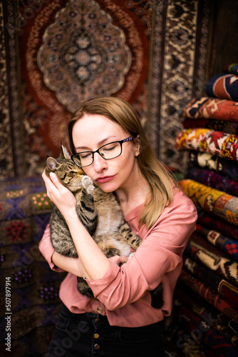 Young happy woman trying to kiss funny turkish cat in oriental  carpet shop. Happy blonde girl in glasses hugging funny fat cat. Woman and cat hugs